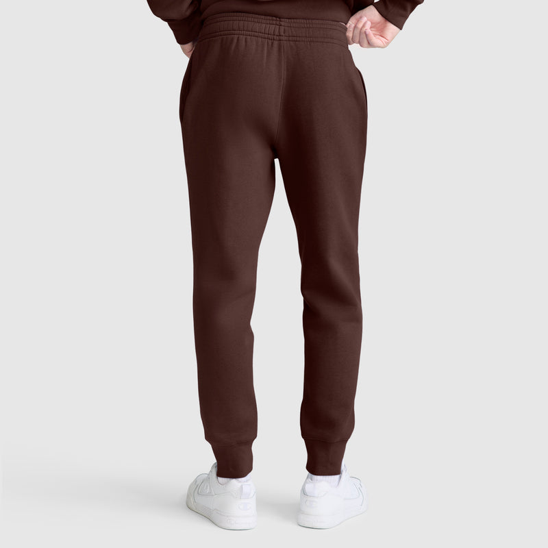 A person is shown from the back wearing Champion Powerblend Joggers, C Logo in brown with white sneakers, standing against a plain light gray background.