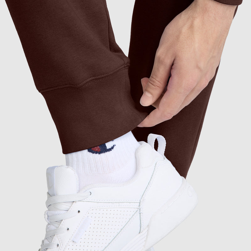 A person adjusts the cuff of Champion Powerblend Joggers featuring the brand’s signature C logo, paired with white sneakers and white socks displaying a small blue and red logo. The background is plain, light grey.