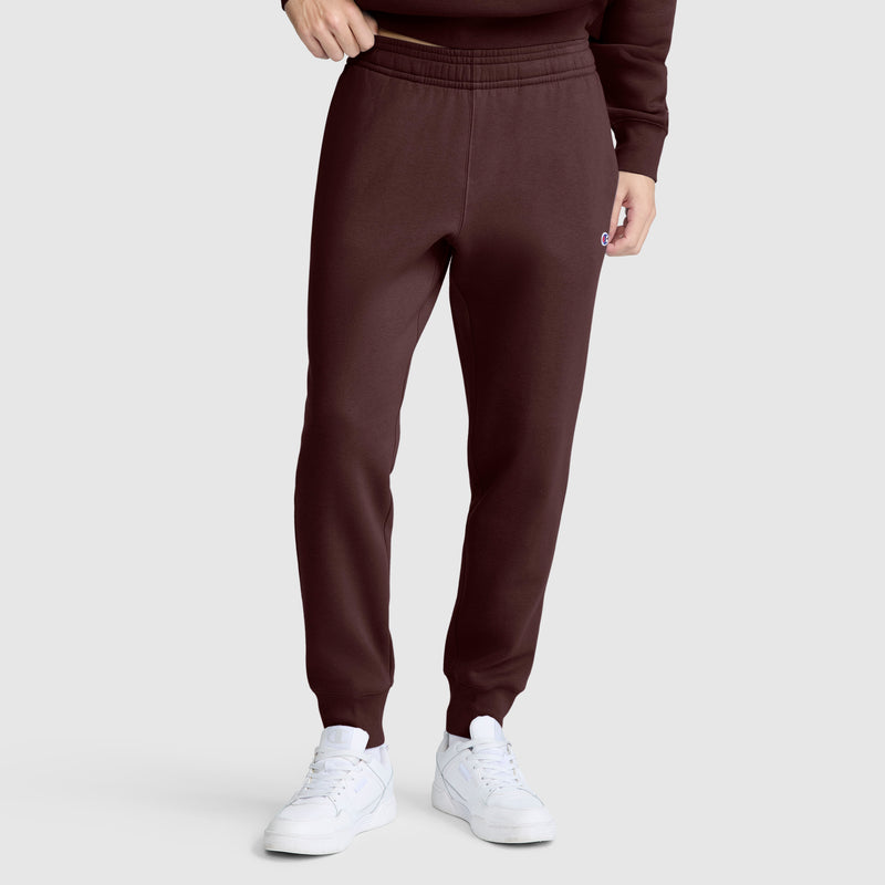 A person is wearing Champion Powerblend Joggers with the C Logo in dark brown, paired with white sneakers and a matching dark brown sweatshirt. Only the lower torso and legs are visible against a plain gray background.