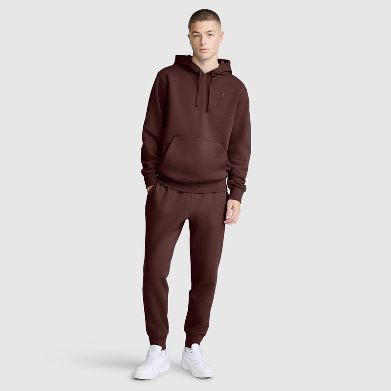 A person stands against a light gray background wearing a matching maroon hoodie and Champion Powerblend Joggers with the C Logo, styled as athletic sweatpants, paired with white sneakers. The outfit is casual, and the person has one hand in their pocket.