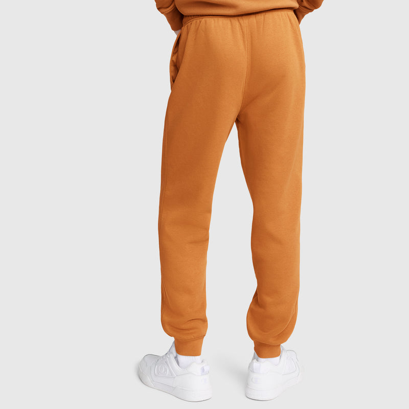 A person is shown from the back against a plain light background, standing with hands in pockets and wearing Champion Powerblend Joggers, C Logo in orange paired with white sneakers.