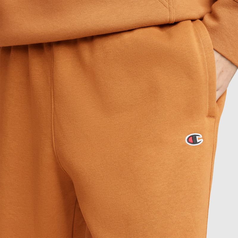 A person wearing Champion Powerblend Joggers, C Logo, in orange with their hand in the pocket; the Champion logo is visible on these men’s joggers.