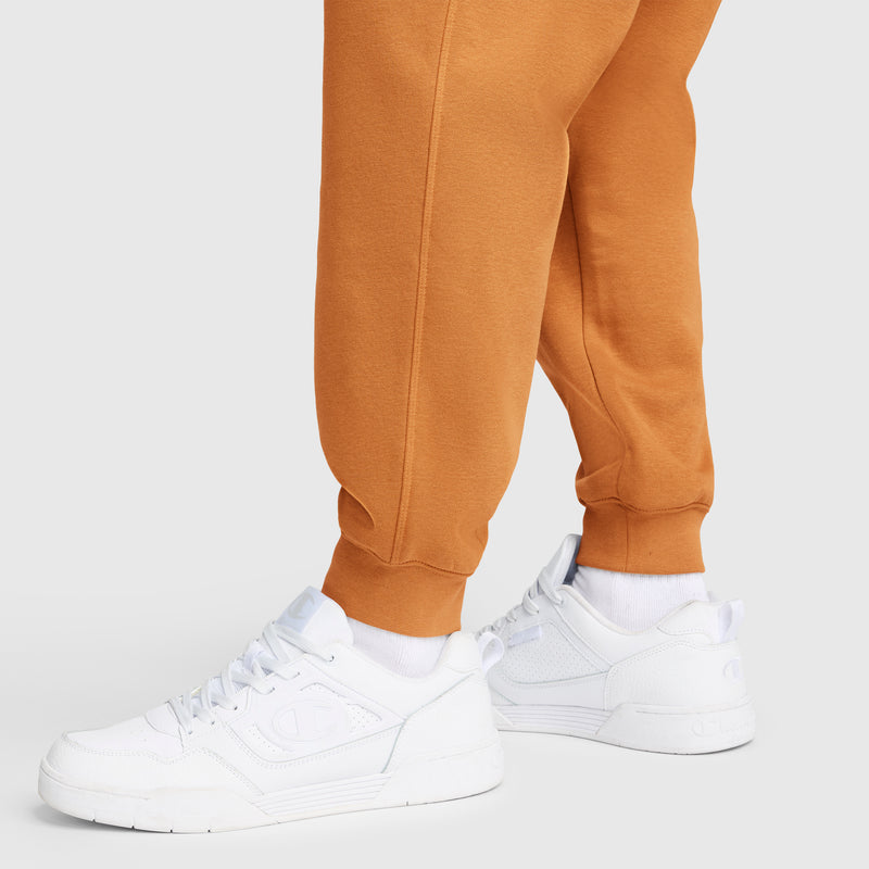 A person wearing Champion Powerblend Joggers, C Logo in orange and white sneakers with white socks stands on a light surface. The image highlights the lower legs and feet, showcasing the athletic sweatpants by Champion and the shoes.