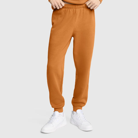 A person wearing Champion Powerblend Joggers, C Logo in orange and white sneakers stands against a plain light gray background. Their hands are adjusting the waistband; only the lower body is visible.
