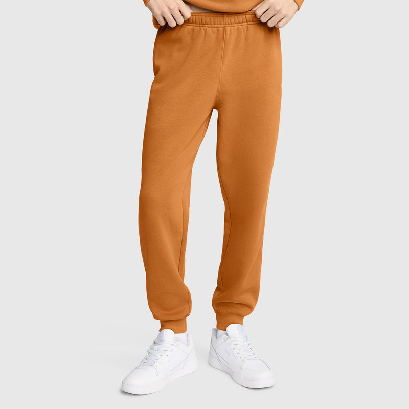 A person wearing Champion Powerblend Joggers, C Logo in orange and white sneakers stands against a plain light gray background. Their hands are adjusting the waistband; only the lower body is visible.