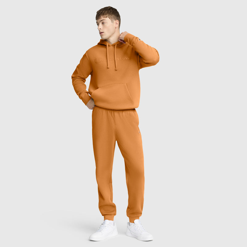 A person stands against a plain background wearing an orange hoodie and Champion Powerblend Joggers, C Logo, paired with white sneakers, looking to the side with one hand on their neck and the other in their pocket.