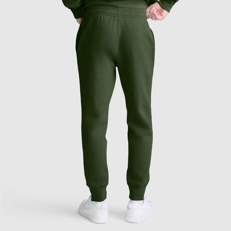A person is shown from the back standing against a plain light gray background, wearing Champion Powerblend Joggers with the C Logo in dark green and white sneakers. Their hands are partially tucked into the waistband.
