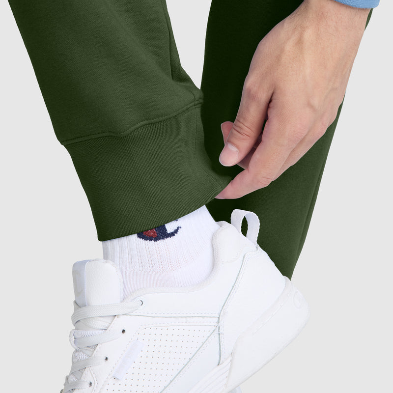 A person wearing green Champion Powerblend Joggers with the C Logo, a blue long sleeve top, white socks, and white sneakers adjusts the cuff of their pants.