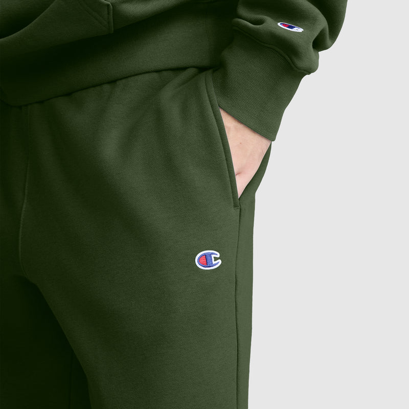 A person wearing dark green Champion Powerblend Joggers, C Logo, paired with a matching sweatshirt, stands with one hand in the pocket. The Champion logo is visible on both the joggers and the sweatshirt sleeve.