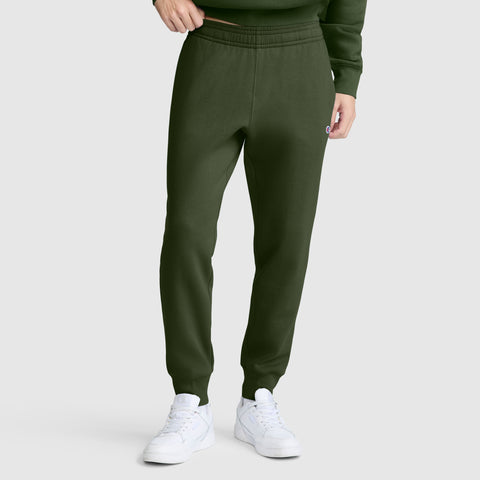 A person wearing dark green Champion Powerblend Joggers with the signature C logo, paired with a matching sweatshirt and white sneakers, standing against a plain light background.