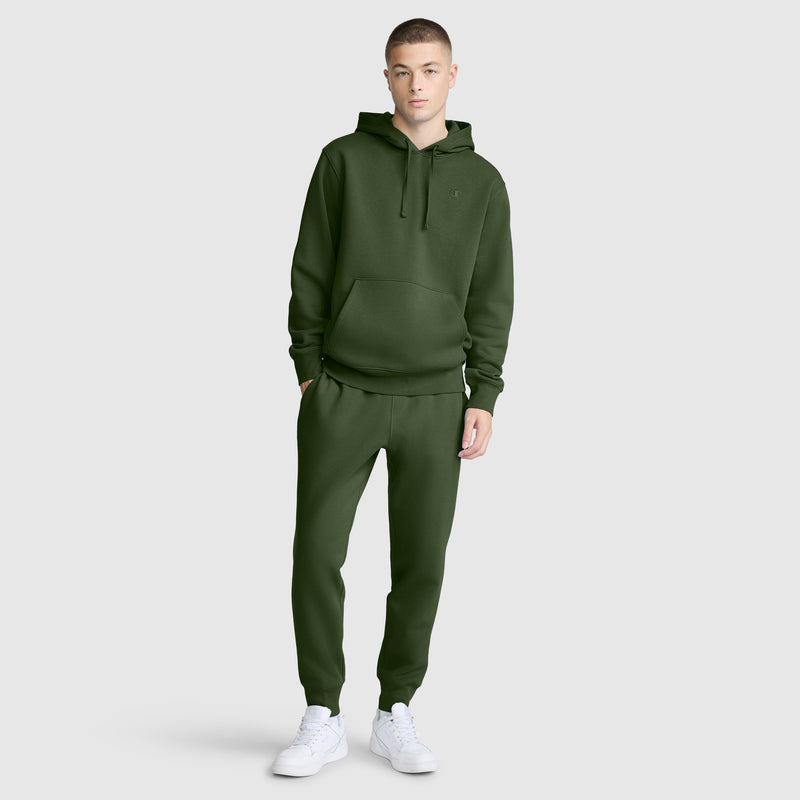 A young man stands against a plain background, wearing a matching dark green hoodie and Champion Powerblend Joggers, C Logo, paired with white sneakers.