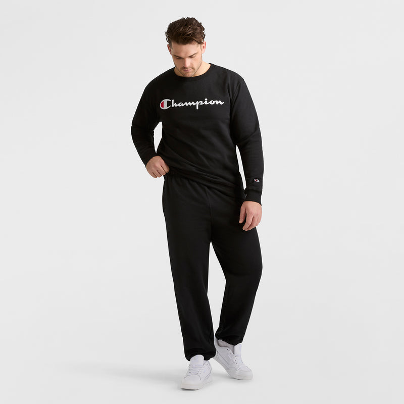 A man wears a black Champion sweatshirt and Champion Powerblend Sweatpants, Cinched Hem, C Logo – Big & Tall. He stands on a white background looking down with one hand in his pocket, finishing the look with white sneakers.