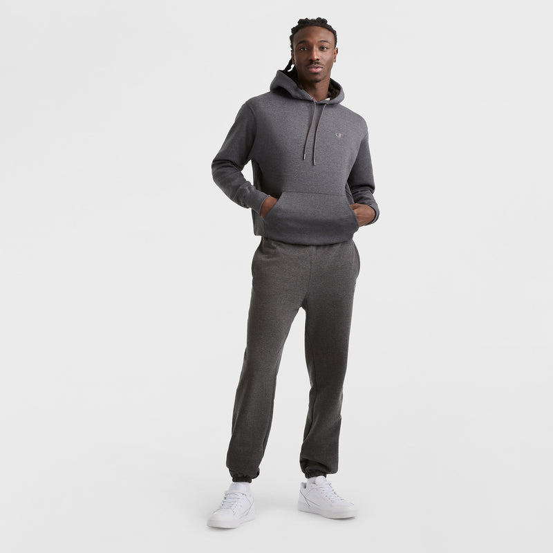 A man stands against a white background wearing Champion Powerblend Sweatpants, Cinched Bottom, C Logo in dark gray with matching hoodie and white sneakers, hands in the hoodie pocket, looking at the camera.