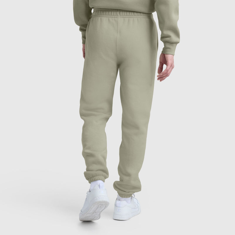 A model wears Champion Powerblend Sweatpants, Cinched Bottom, C Logo in light olive green with a matching sweatshirt, white socks, and sneakers against a plain white background. The pants are made from a soft cotton blend.