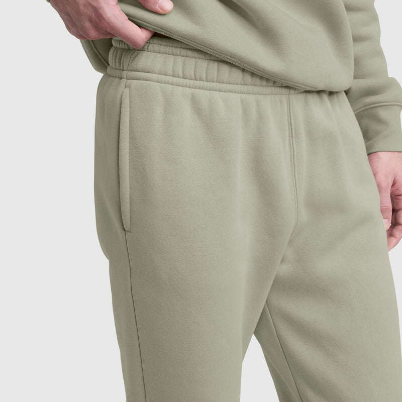 A person wears Champion Powerblend Sweatpants, Cinched Bottom, C Logo in light olive green with a matching sweatshirt. One hand is in the pocket. The image highlights the soft cotton blend fabric and casual style around the waist and upper legs.