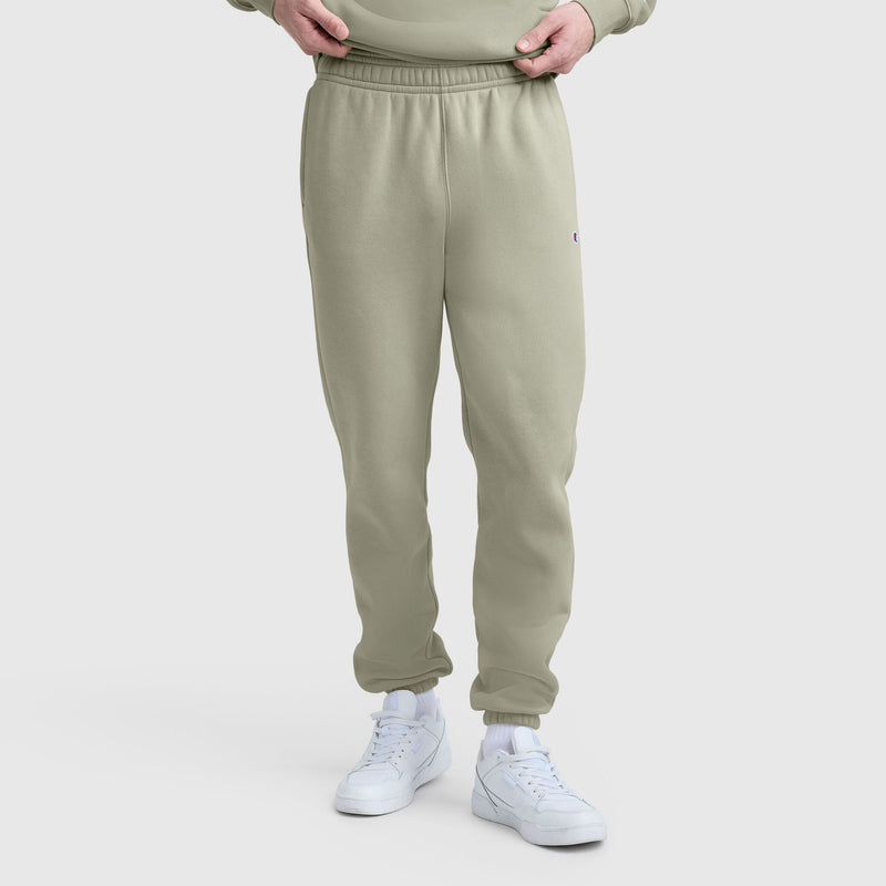 A person wears Champion Powerblend Sweatpants, Cinched Bottom, C Logo in light olive green cotton blend with white sneakers. Only the lower half is visible as hands adjust the waistband against a plain white background.
