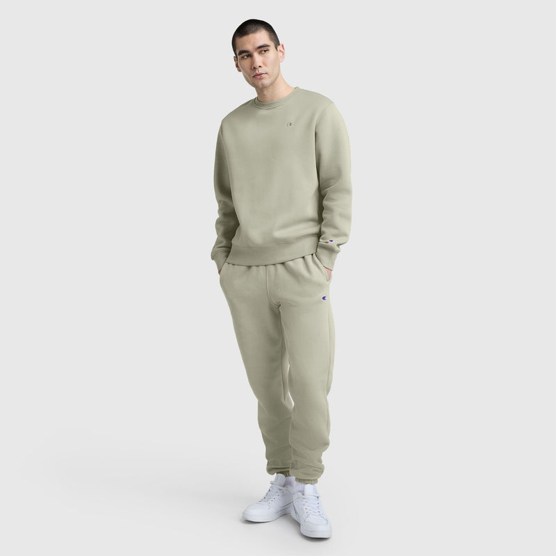 A young man with short dark hair stands against a light gray background, wearing Champion Powerblend Sweatpants, Cinched Bottom, C Logo in light beige with a matching sweatshirt and white sneakers. His hands are in his pockets.