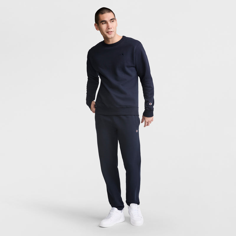 A man stands against a plain white background wearing Champion Powerblend Sweatpants, Cinched Bottom, C Logo in navy blue with a matching sweatshirt and white sneakers, looking slightly to the side with one hand in his pocket.