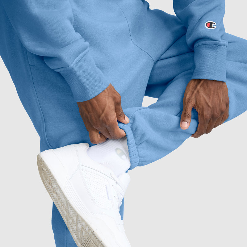 A person wearing light blue Champion Powerblend Sweatpants, Cinched Bottom, C Logo, along with a matching sweatshirt featuring the Champion logo, adjusts the pant leg to reveal white socks and white sneakers against a light background.