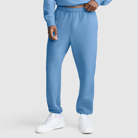 A person wearing Champion Powerblend Cinched Bottom Sweatpants with the C logo in light blue, paired with a matching sweatshirt and white sneakers, stands against a plain light gray background. Only the lower half of their body is visible.