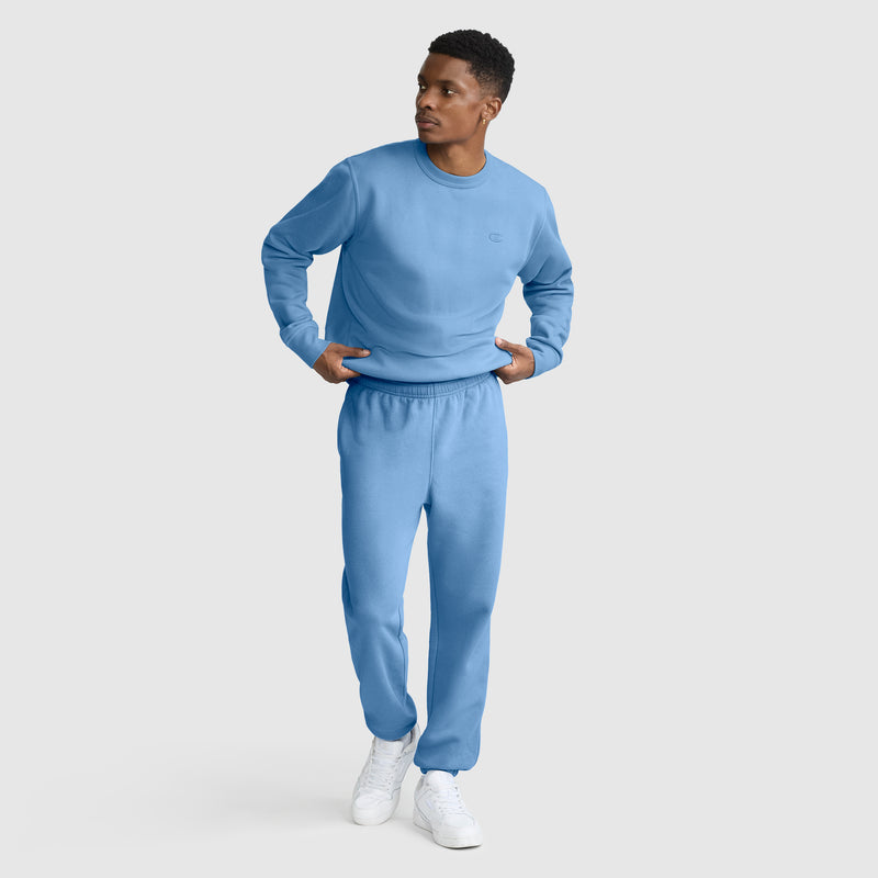A man wearing a light blue sweatshirt and matching Champion Powerblend Sweatpants, Cinched Bottom, C Logo stands with his hands in his pockets, looking to the side. He is also wearing white sneakers. The background is plain and light gray.