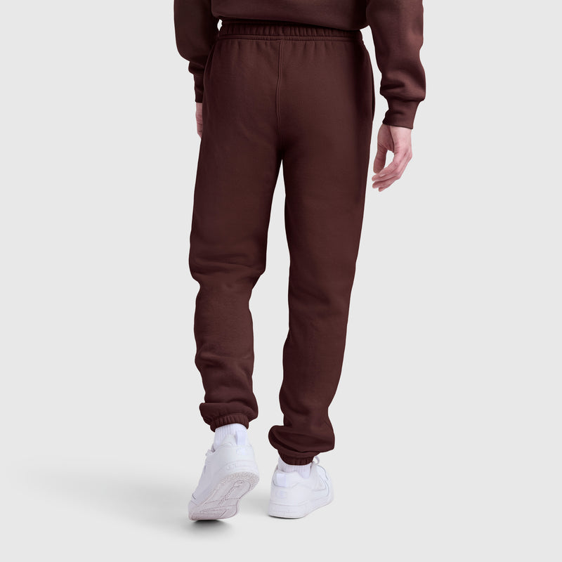 A person wearing Champion Powerblend Sweatpants with a cinched bottom and prominent C logo is shown from the back, standing against a plain light gray background. The brown fleece pants are paired with white sneakers.