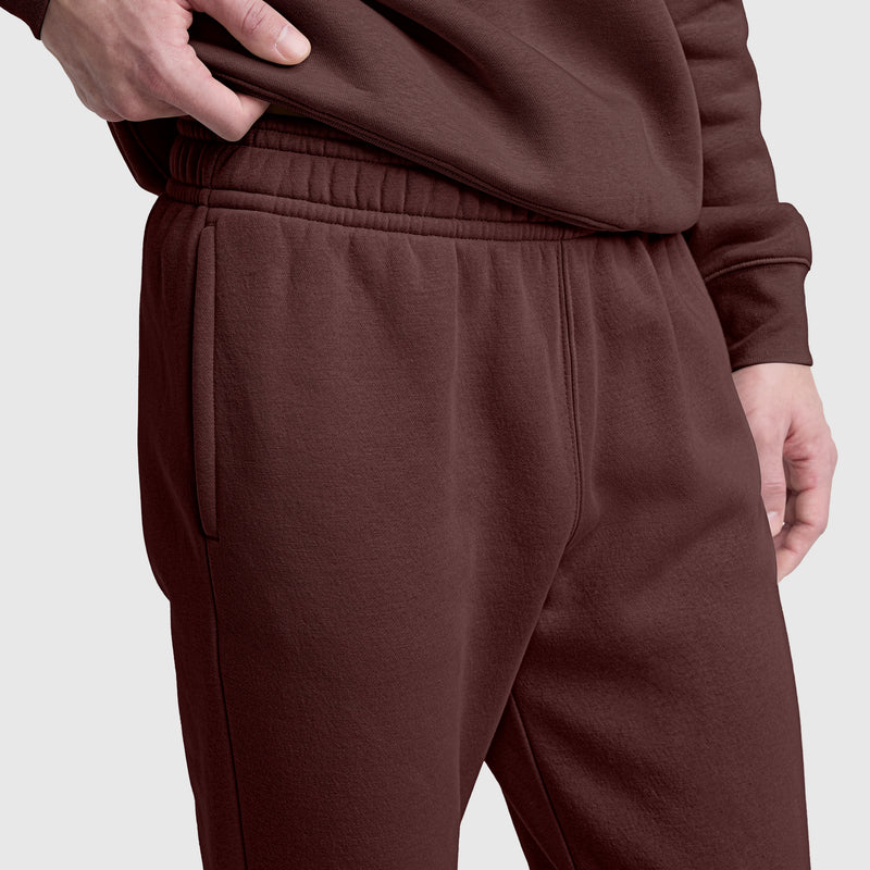 A person stands against a plain light gray background wearing matching brown Champion Powerblend Sweatpants with a cinched bottom and C logo—one hand in their pocket, the other resting by their side.