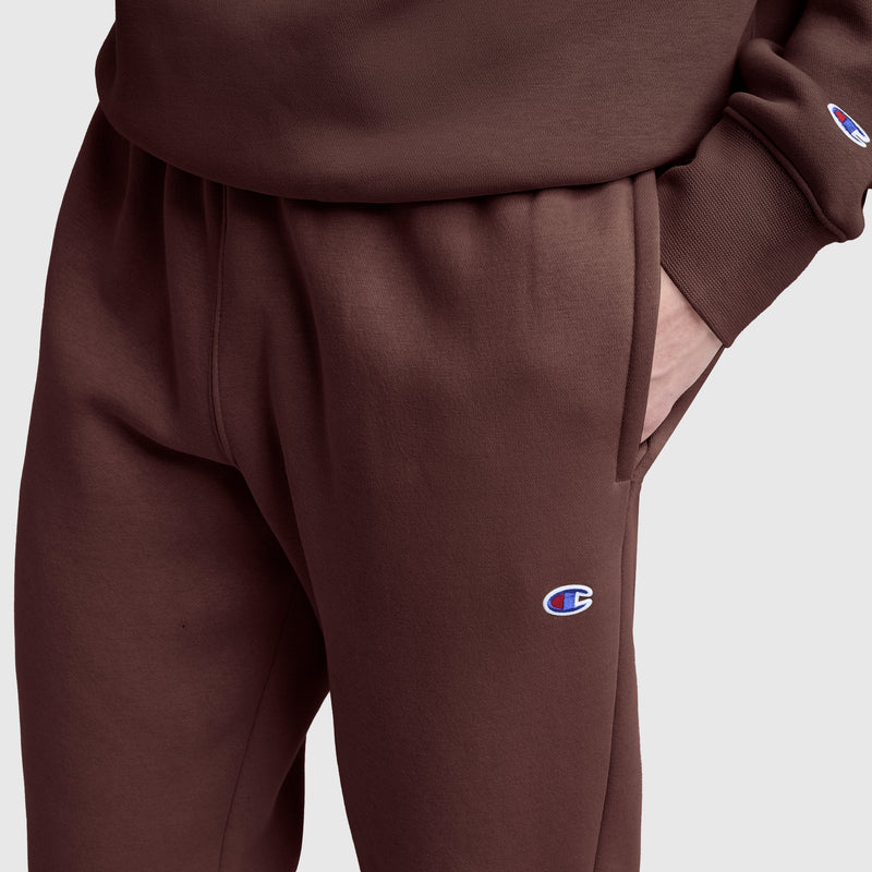 A person is wearing brown Champion Powerblend Sweatpants with a cinched bottom design and the signature C logo, paired with a matching Champion sweatshirt. Their right hand is in the pants pocket, and the Champion logo is visible on both the sleeve and the sweatpants.