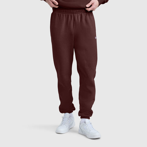 A person is wearing Champion Powerblend Sweatpants, Cinched Bottom, C Logo in burgundy paired with white sneakers, standing against a plain light gray background. Only the lower half of the body is visible, with hands tucked into the waistband. The cinched bottom design of these Champion sweatpants offers a comfortable and modern look.