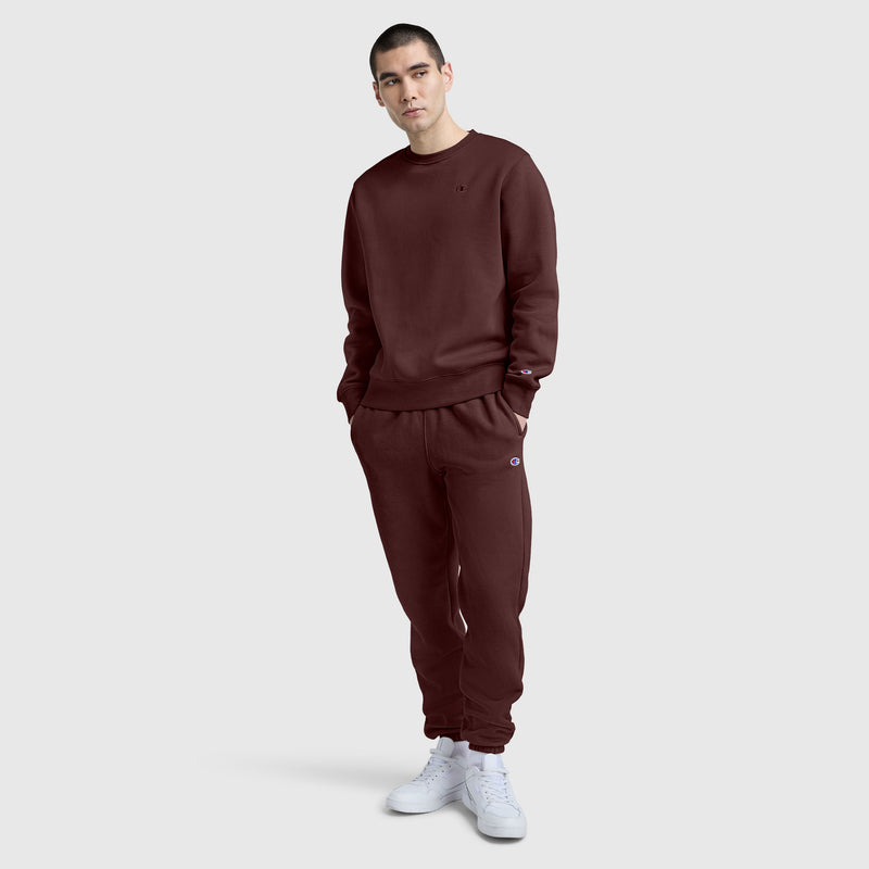 A person stands against a plain background wearing a matching maroon sweatshirt and Champion Powerblend Sweatpants with Cinched Bottom and C Logo, paired with white sneakers, looking at the camera with hands in their pockets.
