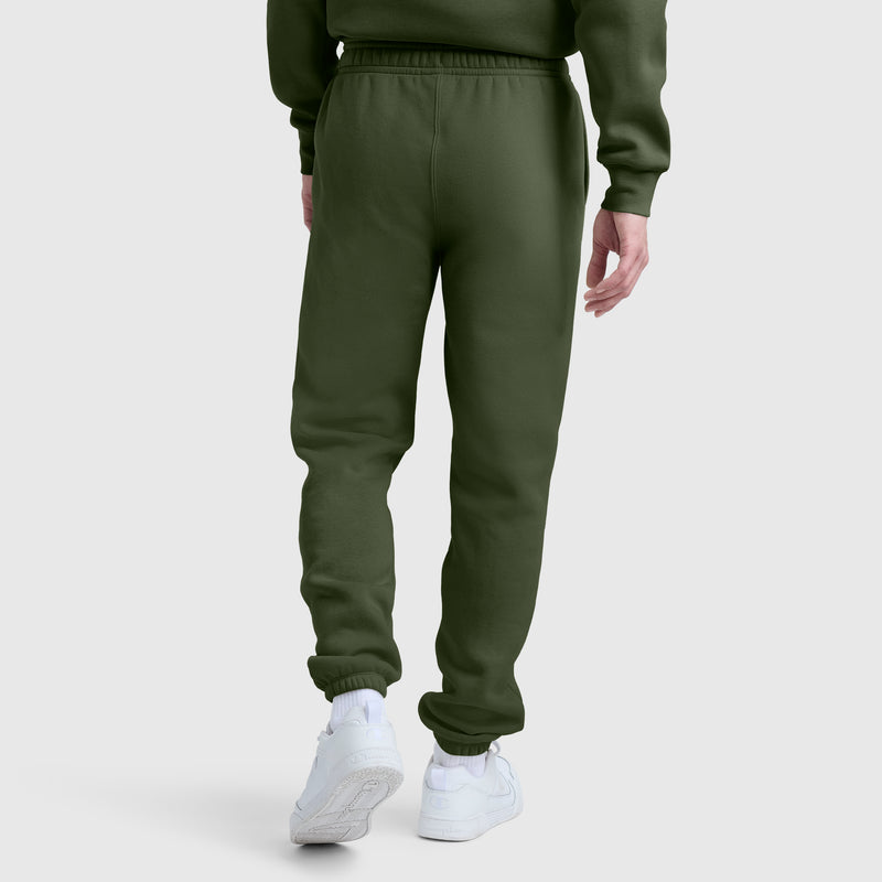 A man is shown from the back wearing olive green Champion Powerblend Sweatpants with a cinched bottom and matching sweatshirt, standing on a white surface and pairing the look with white sneakers. These men’s fleece pants feature Champion’s signature C logo and offer a comfortable, modern athletic style.