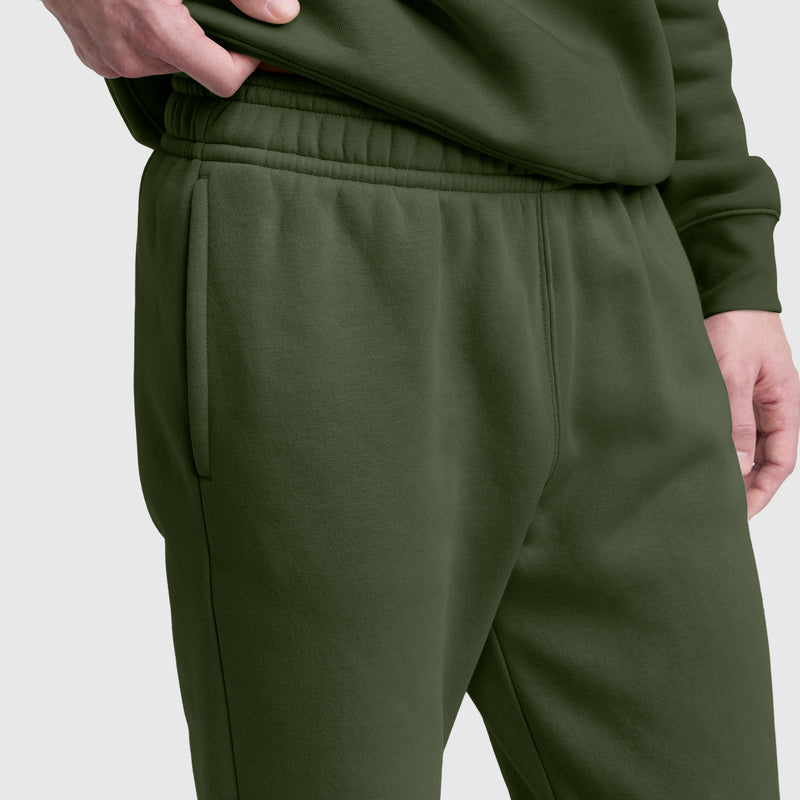 A person wearing dark green Champion Powerblend Sweatpants with a cinched bottom and C Logo, paired with a matching sweatshirt, stands with one hand resting on their hip. The sweatpants feature an elastic waistband, convenient side pockets, and the signature Powerblend cinched cuffs for added comfort and style.