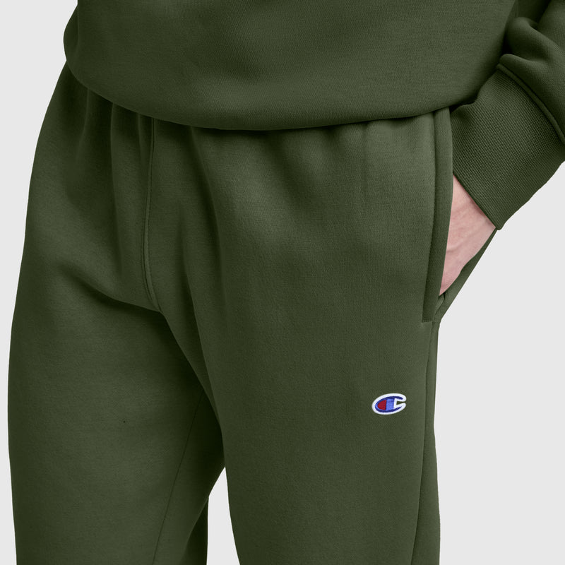 A person is wearing olive green Champion Powerblend Sweatpants with a cinched bottom and the iconic C logo on the left thigh, paired with a matching sweatshirt and one hand in their pocket. The background is light gray.