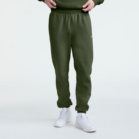 A person is wearing Champion Powerblend Sweatpants with a cinched bottom and C logo in olive green, paired with white sneakers, standing against a plain white background. Only the lower half of the body is visible.