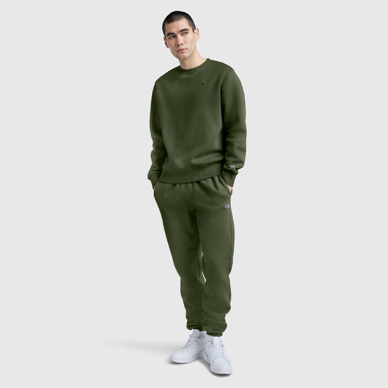 A person stands against a plain light gray background, wearing a matching olive green sweatshirt and Champion Powerblend Sweatpants with Cinched Bottom and C Logo, paired with white sneakers.