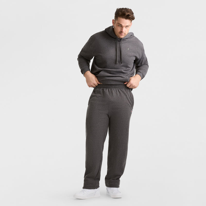 A man stands against a white background wearing matching dark gray Champion Powerblend Sweatpants, Open Hem, C Logo - Big & Tall, a hoodie, and white sneakers.