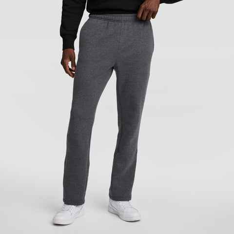 A person wearing Champion Powerblend Sweatpants, Open Hem, C Logo in charcoal gray stands against a plain light gray background, paired with a black sweatshirt and white sneakers, left hand resting on their waistband.