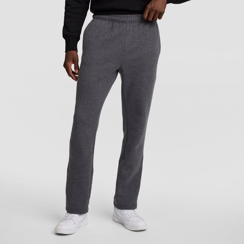 A person wearing Champion Powerblend Sweatpants, Open Hem, C Logo in charcoal gray stands against a plain light gray background, paired with a black sweatshirt and white sneakers, left hand resting on their waistband.