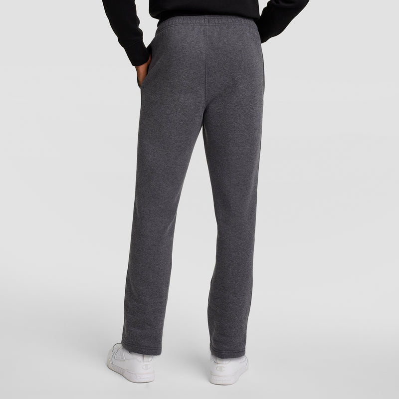 Someone is shown from the back with hands in pockets, wearing dark gray Champion Powerblend Sweatpants, Open Hem, C Logo, a black top, and white sneakers against a plain light background.