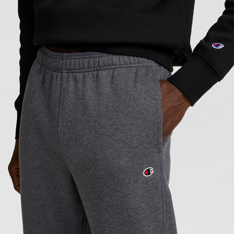 A person wears a black sweatshirt and gray Champion Powerblend Sweatpants, Open Hem, C Logo, with one hand in their pocket. Both the sweatshirt cuff and the sweatpants show the Champion logo.