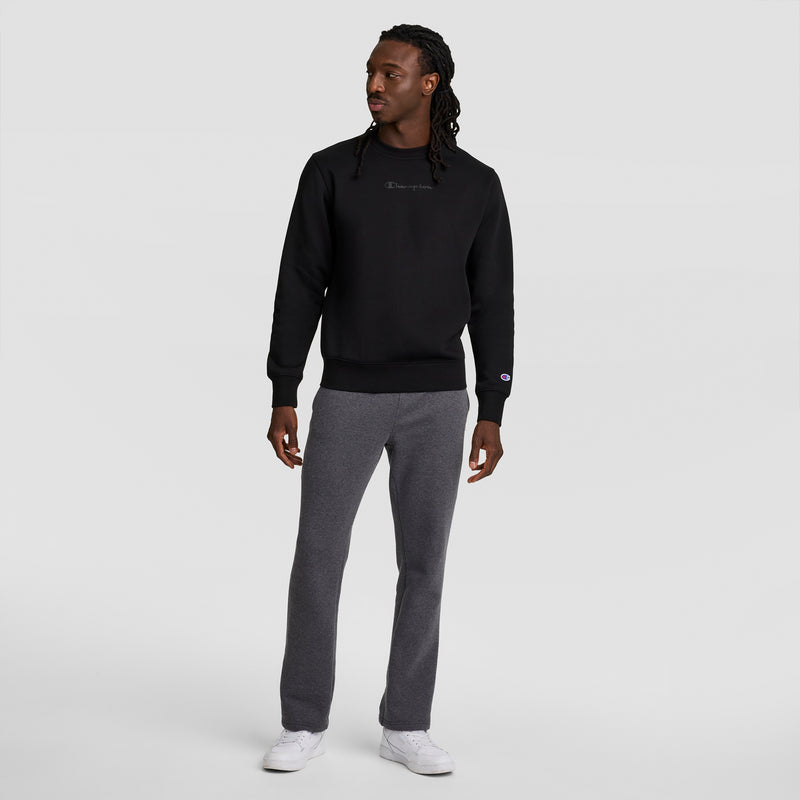 A man faces slightly left, wearing a black sweatshirt, Champion Powerblend Sweatpants with an open hem and C Logo, plus white sneakers, against a plain light gray background.