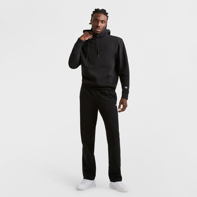 Against a plain white background, a person wears Champion Powerblend Sweatpants, Open Hem, C Logo, paired with a black hoodie and white sneakers. They raise one hand to their chin and look at the camera.