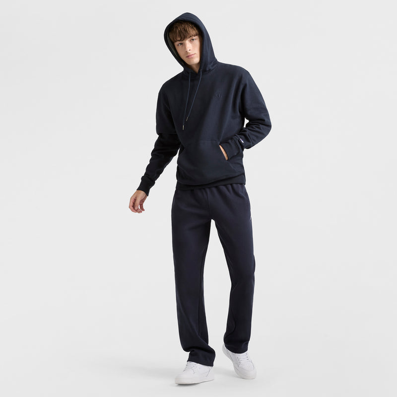 A person wearing Champion Powerblend Sweatpants, Open Hem, C Logo, and a dark hooded sweatshirt with white sneakers stands against a plain white background.