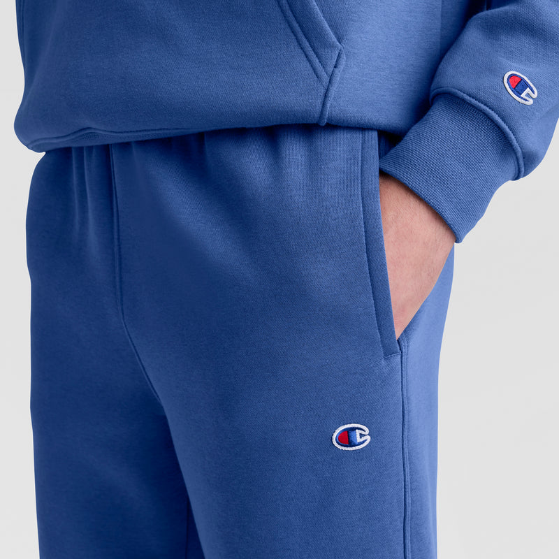 A person wearing matching blue Champion Powerblend Sweatpants, Open Hem, C Logo and sweatshirt, with one hand in their pocket. The Champion logo appears on both the sleeve and pant leg.