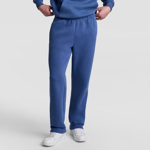 A person wearing Champion Powerblend Sweatpants, Open Hem, C Logo in blue stands against a plain light gray background. The image is cropped at the chest, displaying only the lower torso and legs.