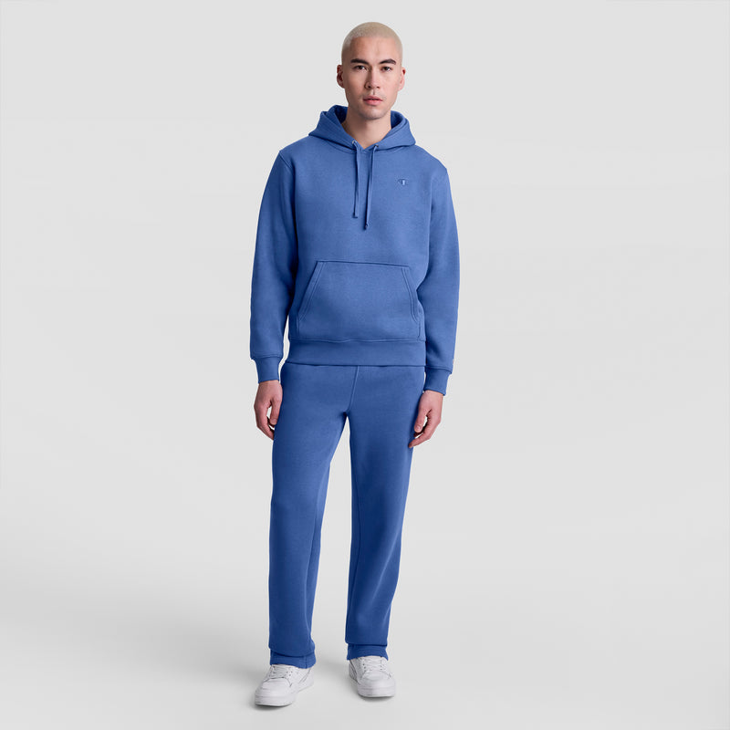 A person with short blond hair faces forward against a light gray backdrop, wearing a matching blue hoodie and Champion Powerblend Sweatpants, Open Hem, C Logo, paired with white sneakers.