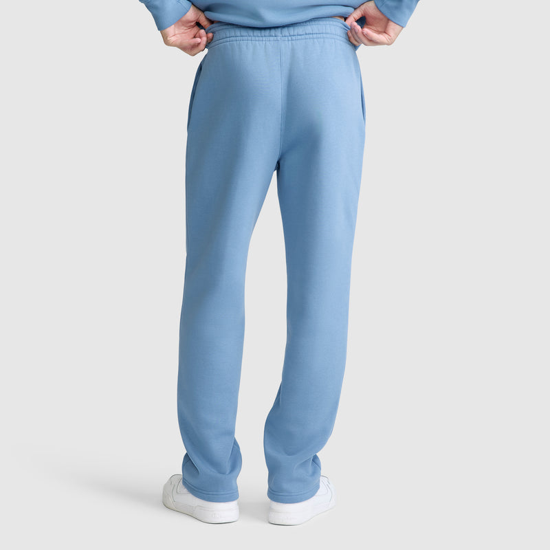 A person is seen from behind, standing with hands on hips and wearing Champion Powerblend Sweatpants, Relaxed Bottom, C Logo in light blue along with white sneakers. The image highlights the back view of the pants against a plain light background.