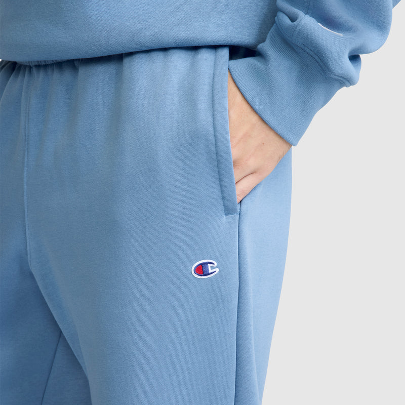 A person wears light blue Champion Powerblend Sweatpants, Relaxed Bottom, C Logo, paired with a matching sweatshirt and has one hand in their pocket. The Champion logo is embroidered near the pocket on the sweatpants.