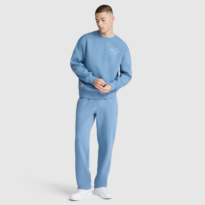 A young man stands with his hands together, looking down, dressed in a light blue Champion Powerblend Sweatpants with a relaxed bottom and C logo. He is also wearing matching apparel and white sneakers against a plain light gray background.