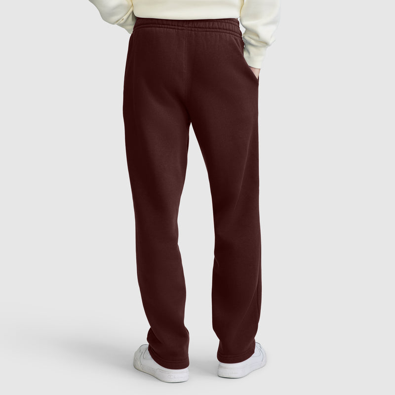 A person is shown from the back with hands in pockets, standing against a plain light gray background, wearing maroon Champion Powerblend Sweatpants with a relaxed bottom and C logo. The open-hem sweatpants provide everyday comfort and are paired with white sneakers and a cream sweatshirt.